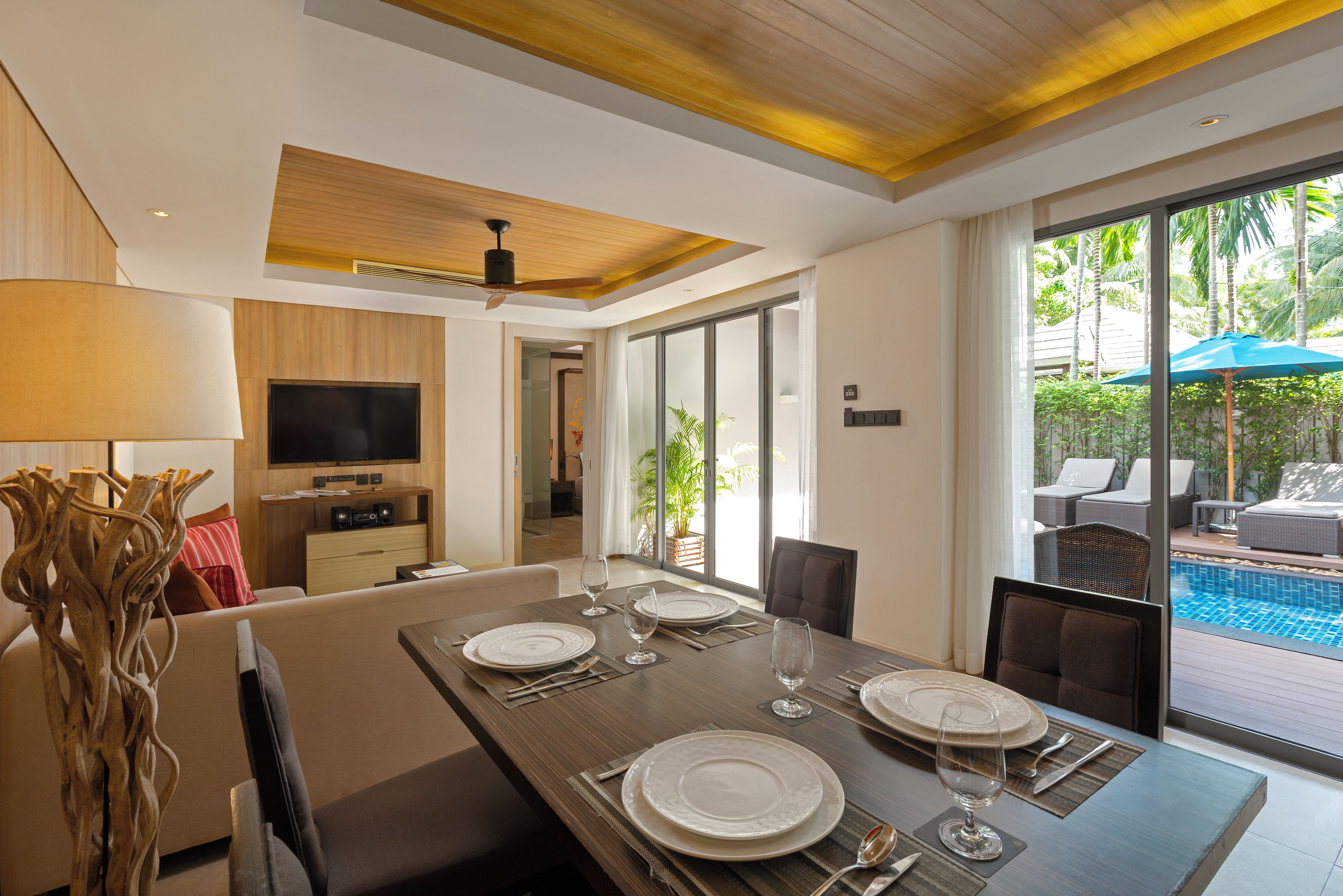 Comedor con mesa preparada para cuatro, televisor y vista a una piscina exterior.