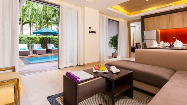 A modern hotel room with a sofa, a small table with a fruit bowl, a view of a pool with lounge chairs, and a red accent wall.