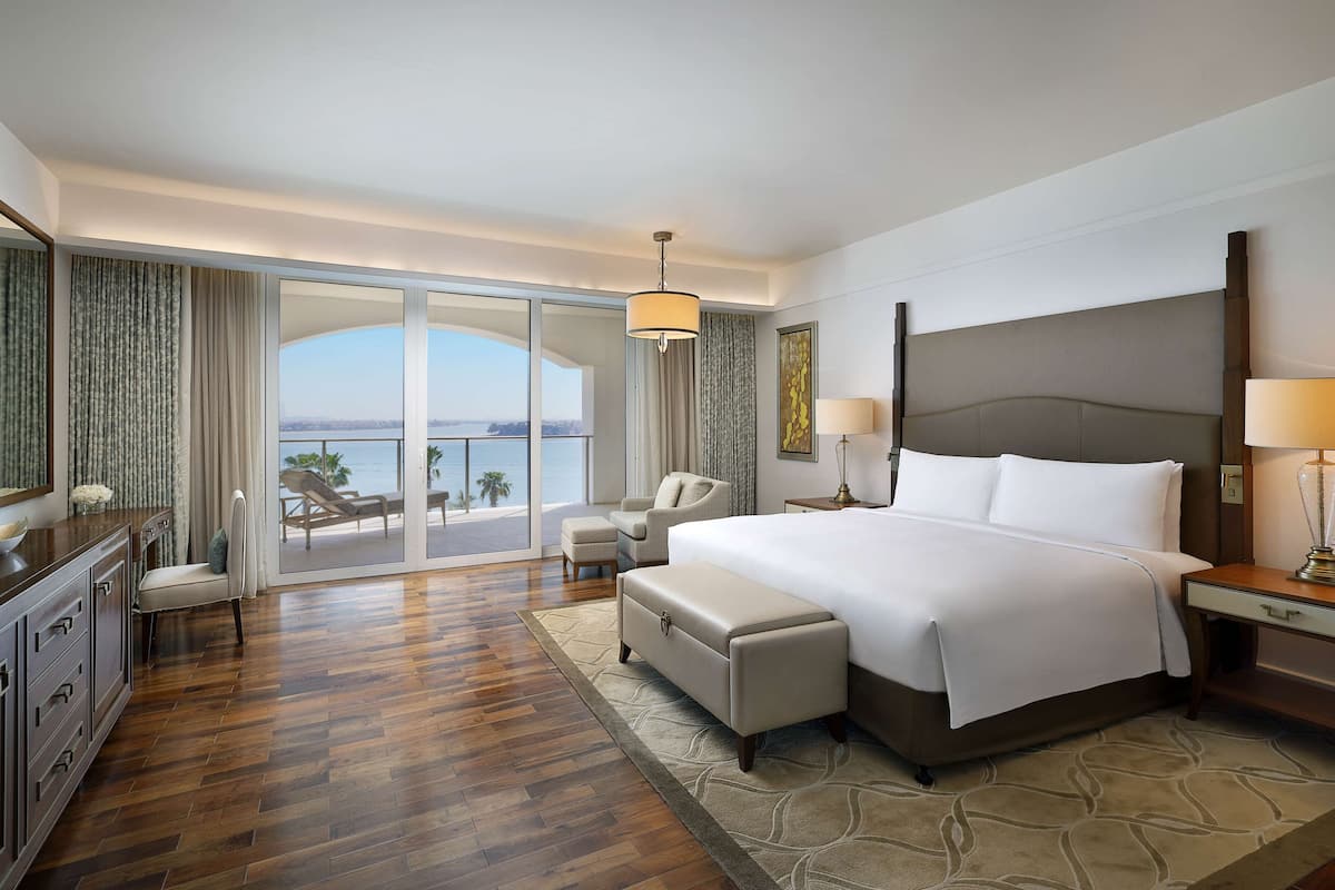 A spacious hotel room with a large bed, a desk, a chair, and a view of the ocean through a sliding glass door.