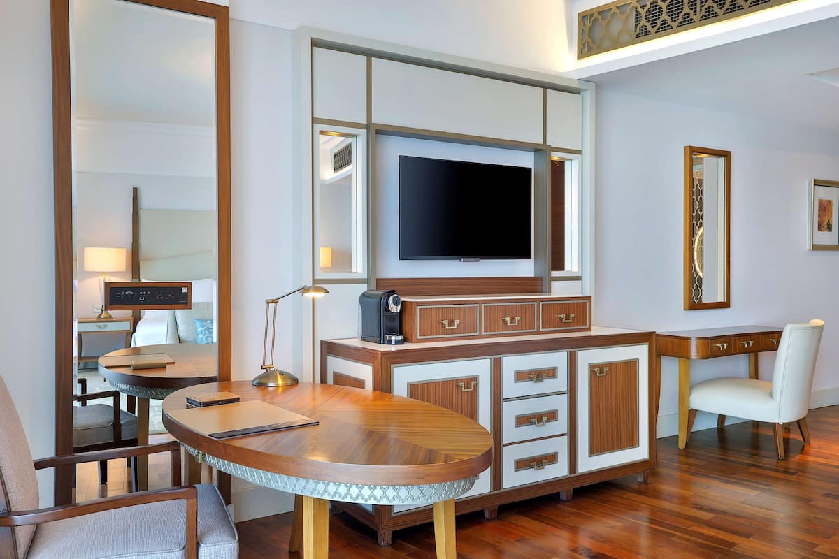 A modern hotel room with a wooden dining table, a flat-screen TV mounted on the wall, a minibar, and a seating area with a chair.