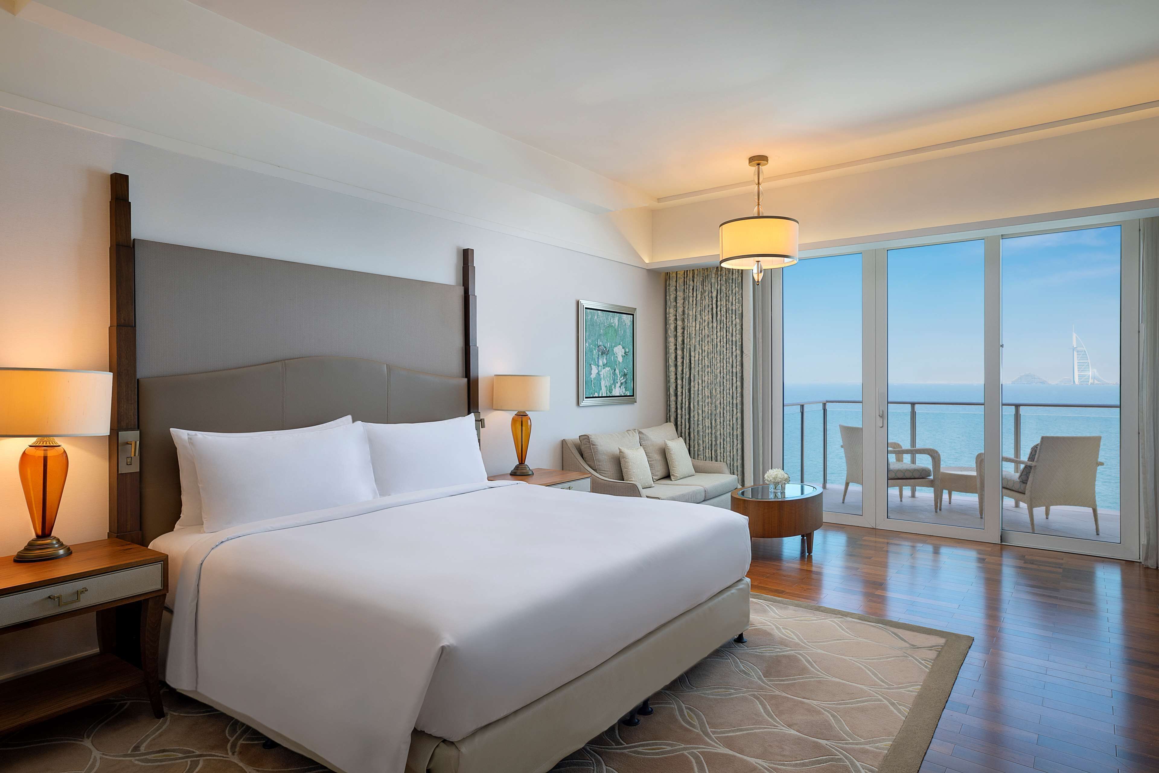 A hotel room with a large bed, a sitting area with a view, and a balcony with a view of the sea.