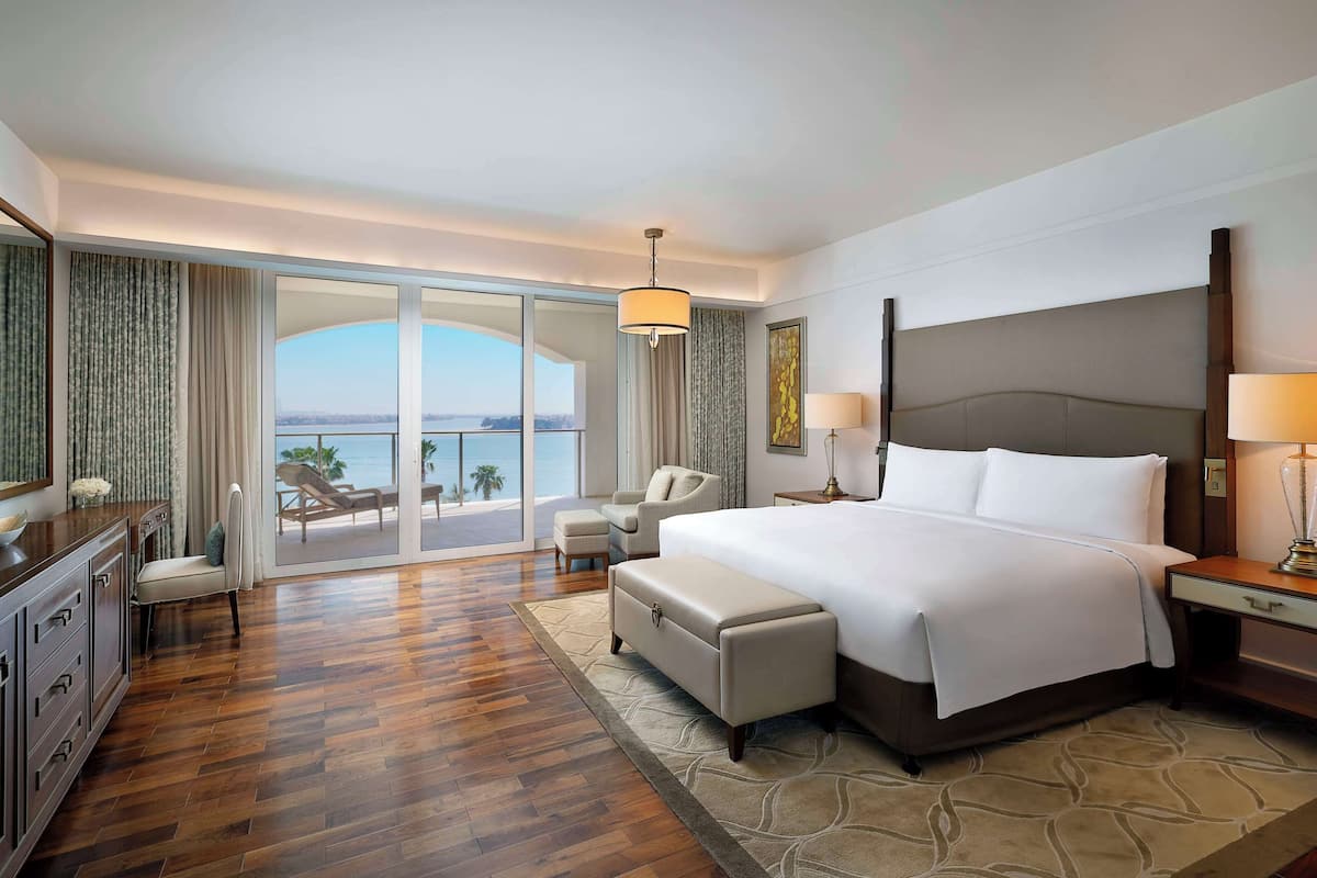 A hotel room with a large bed, a desk, a chair, a small table, and a view of the ocean.