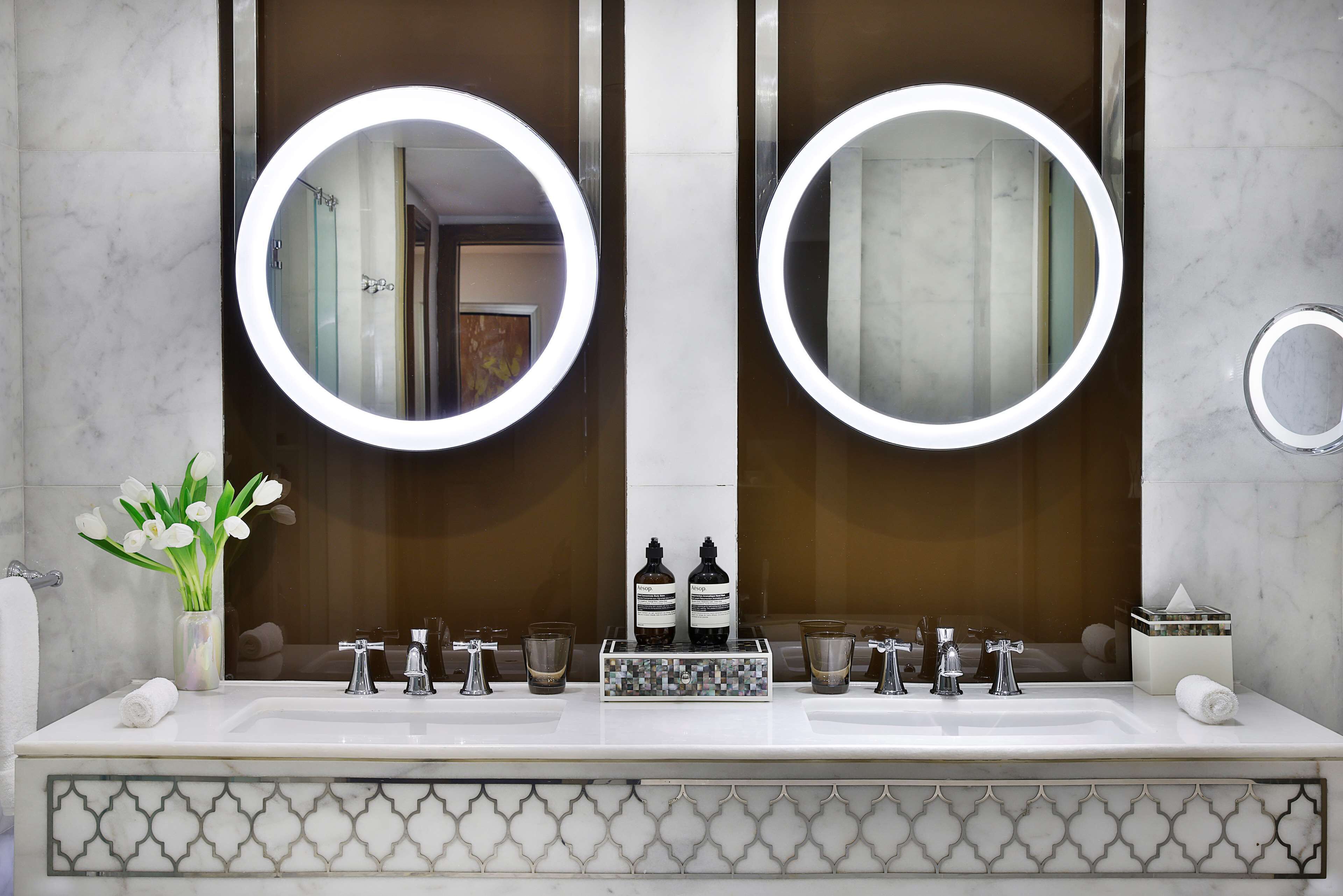 Un baño con dos lavamanos, un espejo grande con luces redondas, un florero y un detalle decorativo en los azulejos.
