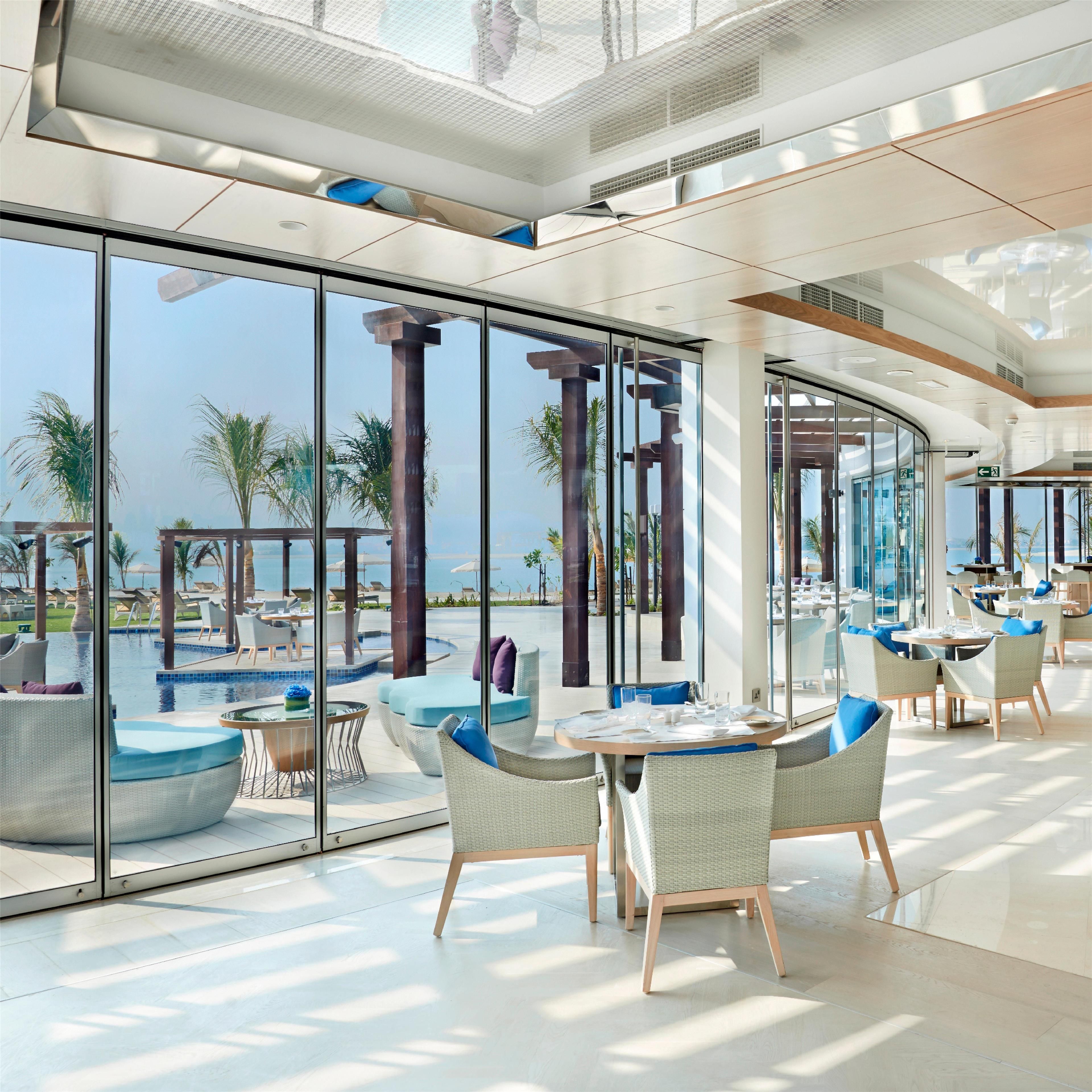 A modern hotel lobby with large glass windows, a pool area, and outdoor seating.