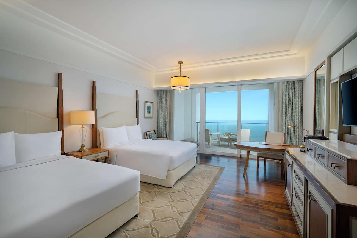 A hotel room with two beds, a desk, a chair, a TV, and a balcony with a view of the sea.