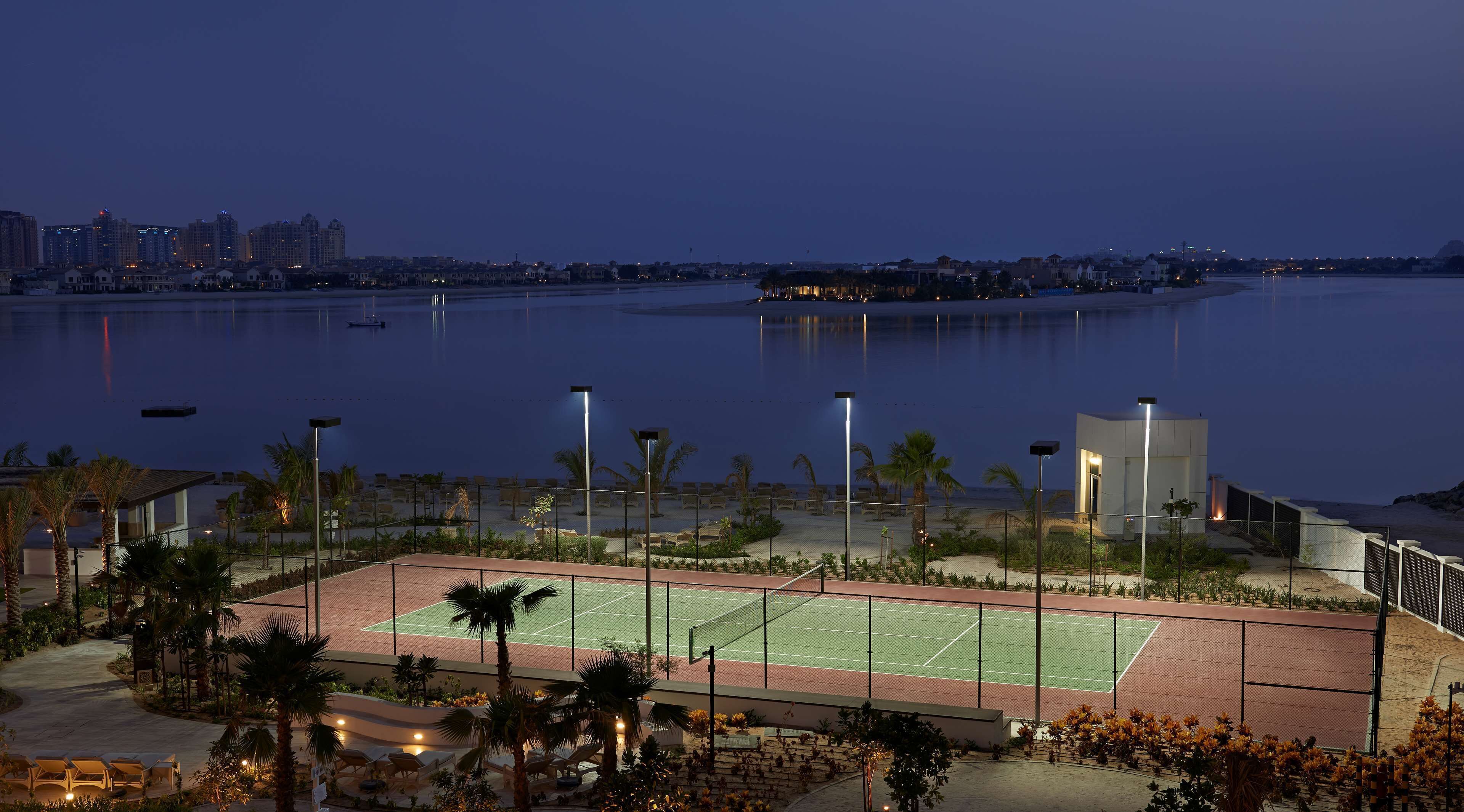 A tennis court with surrounding palm trees and a view of a city skyline at dusk.