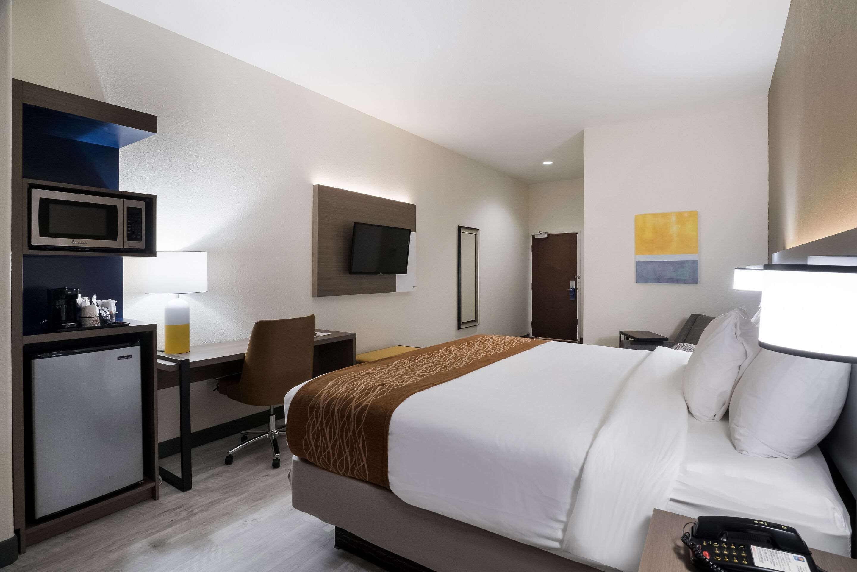 Habitación de hotel con cama, escritorio, silla, microondas, refrigerador, televisor y obras de arte en las paredes.