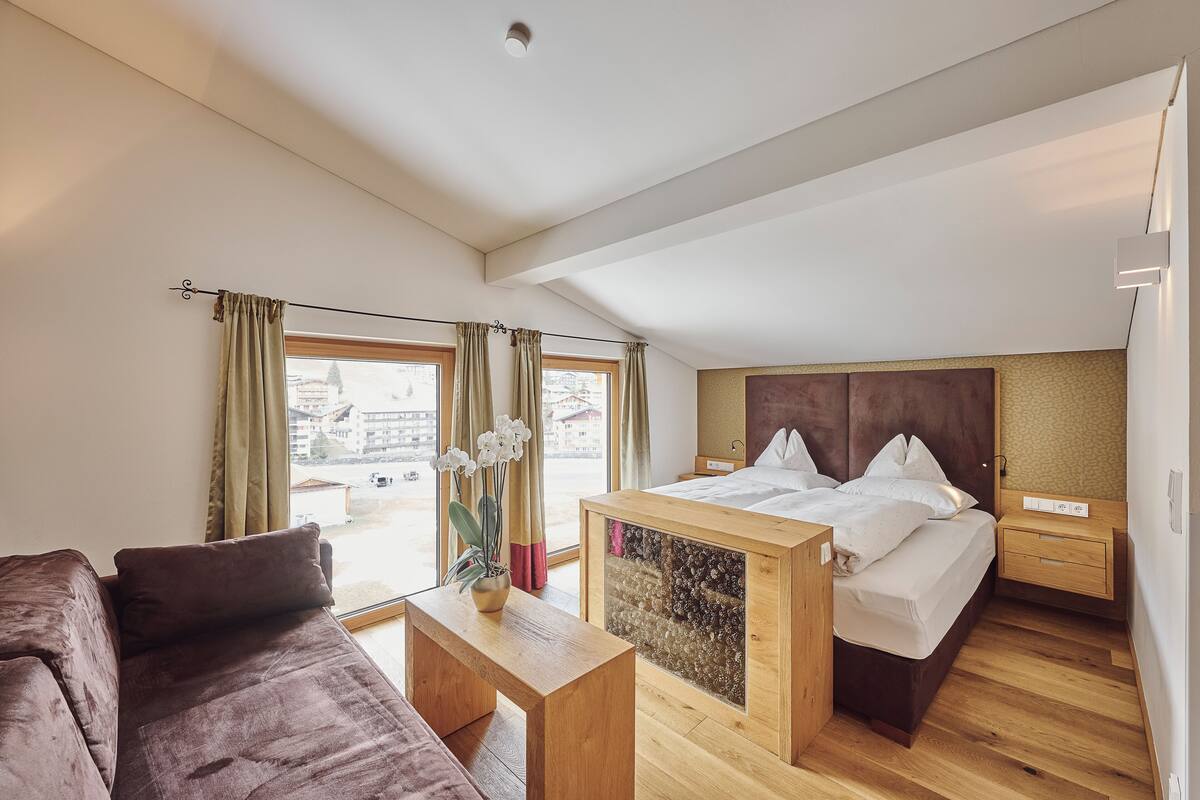 A modern bedroom with a large bed, a wooden bedside table, a nightstand, and a view of the city through large windows.