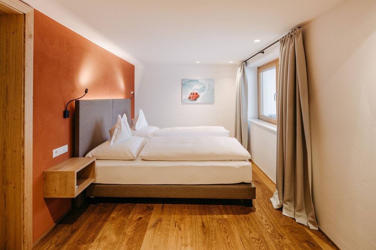 A modern bedroom with a wooden floor, a bed with white linens, a nightstand, a wall-mounted lamp, a window with curtains, and a painting on the wall.