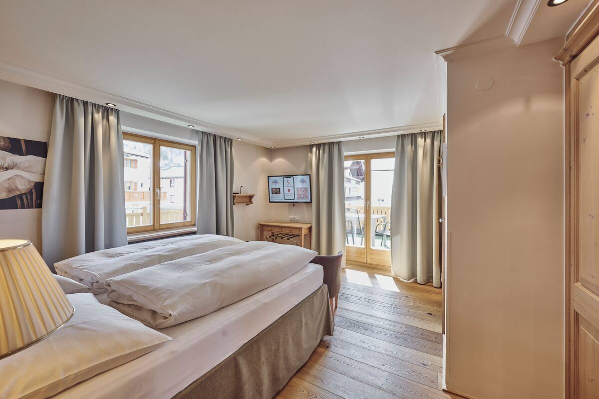 A bedroom with a bed, a desk, and a TV. There is a balcony with curtains.