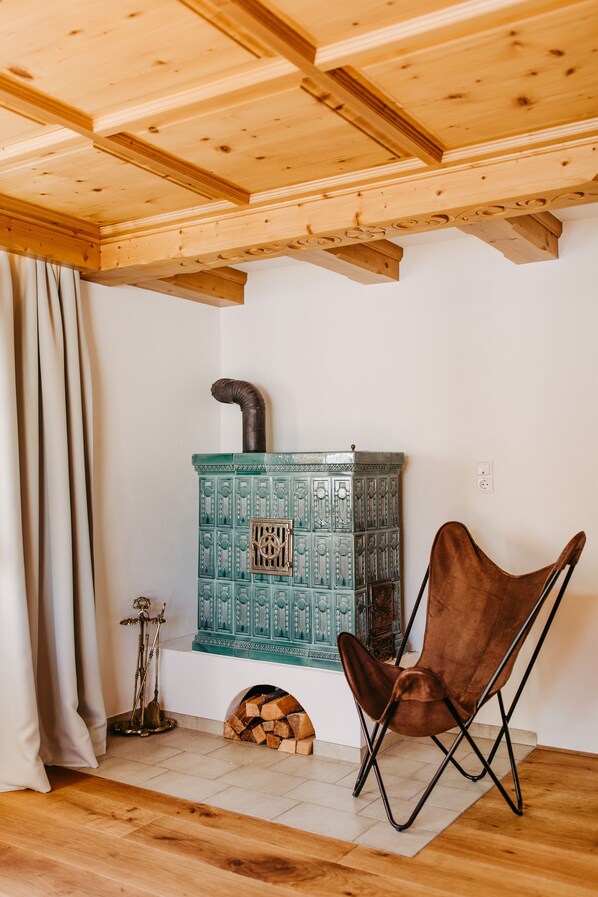 A room with a wooden ceiling, a blue patterned stove, a brown chair, and a curtain. - Fernsicht Alpen-Apartments (Lech am Arlberg)