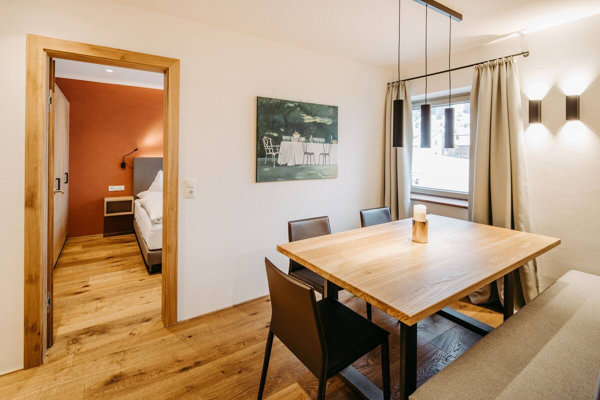 A hotel room with a wooden dining table, chairs, a bed, a painting, and a window with curtains.