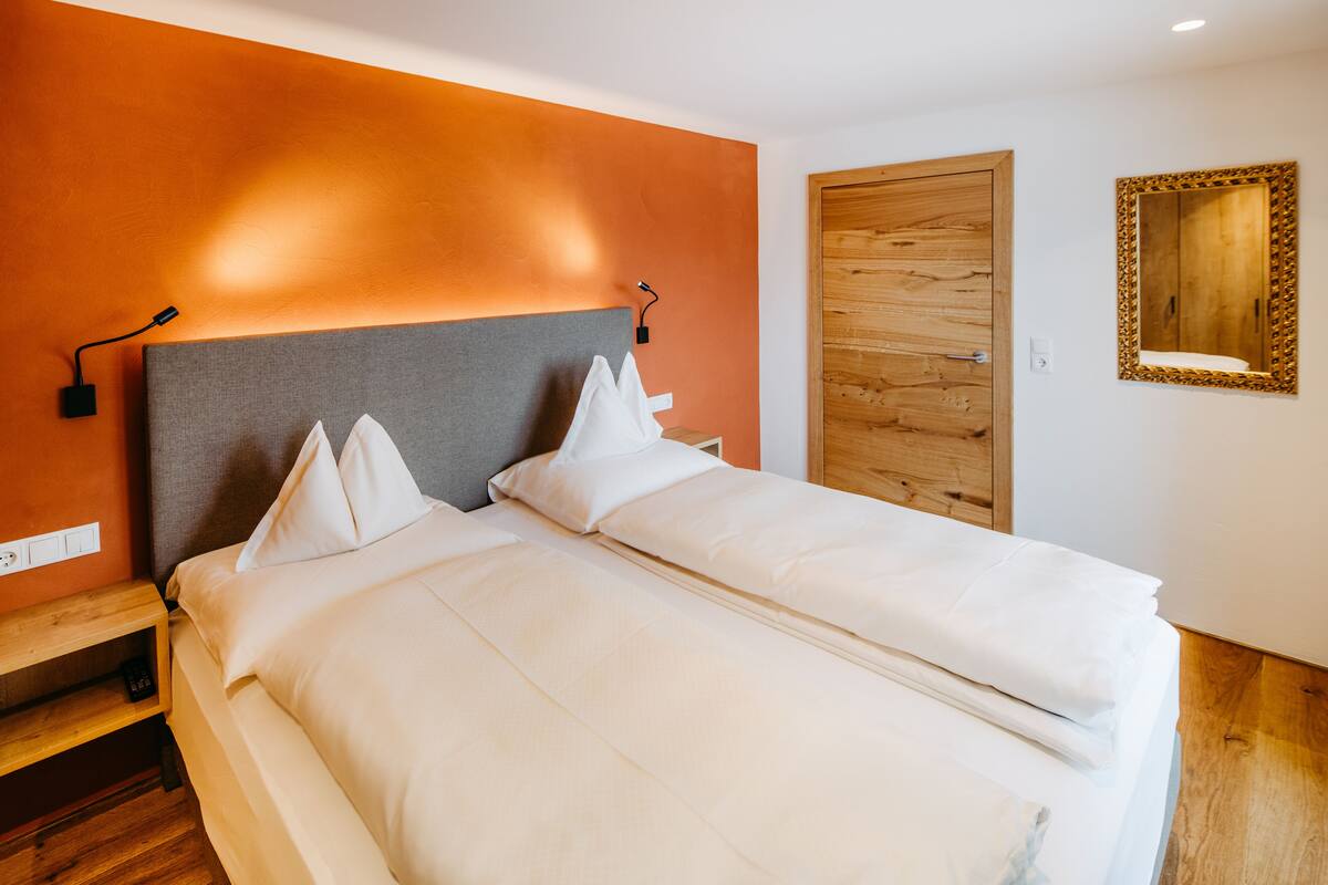 A double bed with white linens and a grey headboard, a wooden nightstand, and a wooden door with a mirror.