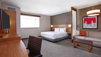 A hotel room with a bed, a desk, a chair, a sofa, a coffee table, a TV, a window, and a framed picture on the wall.