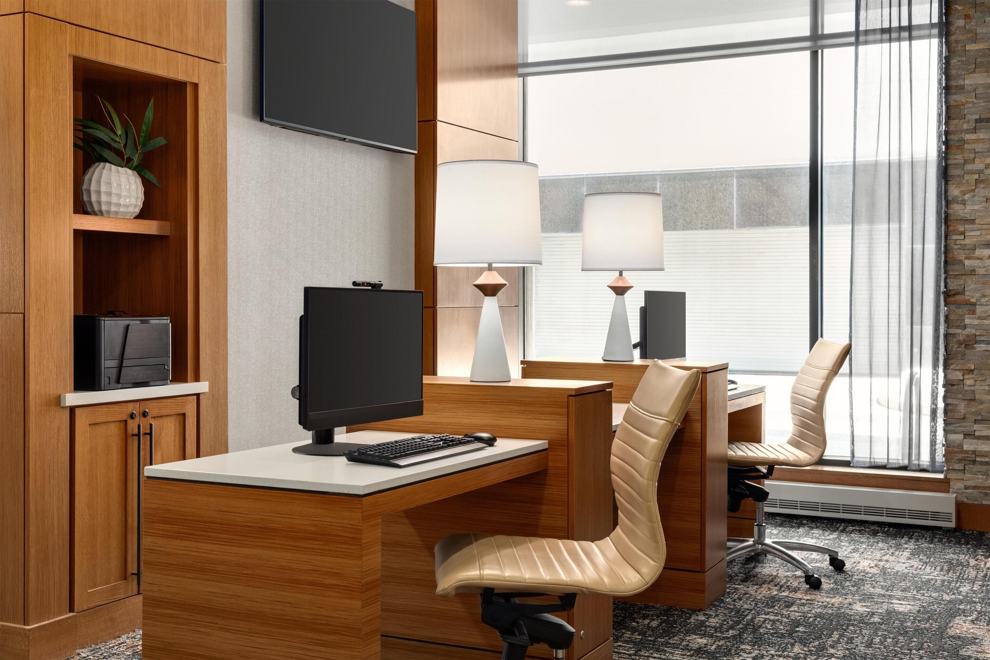 Quarto de hotel com escrivaninha de madeira, um computador, uma cadeira e uma janela grande com persianas.