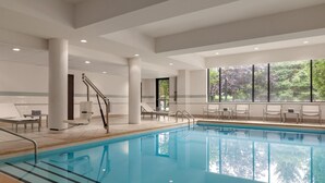An indoor swimming pool with clear blue water, surrounded by white walls and columns, with a view of trees through large windows. - Hyatt Place Bloomington (Bloomington)
