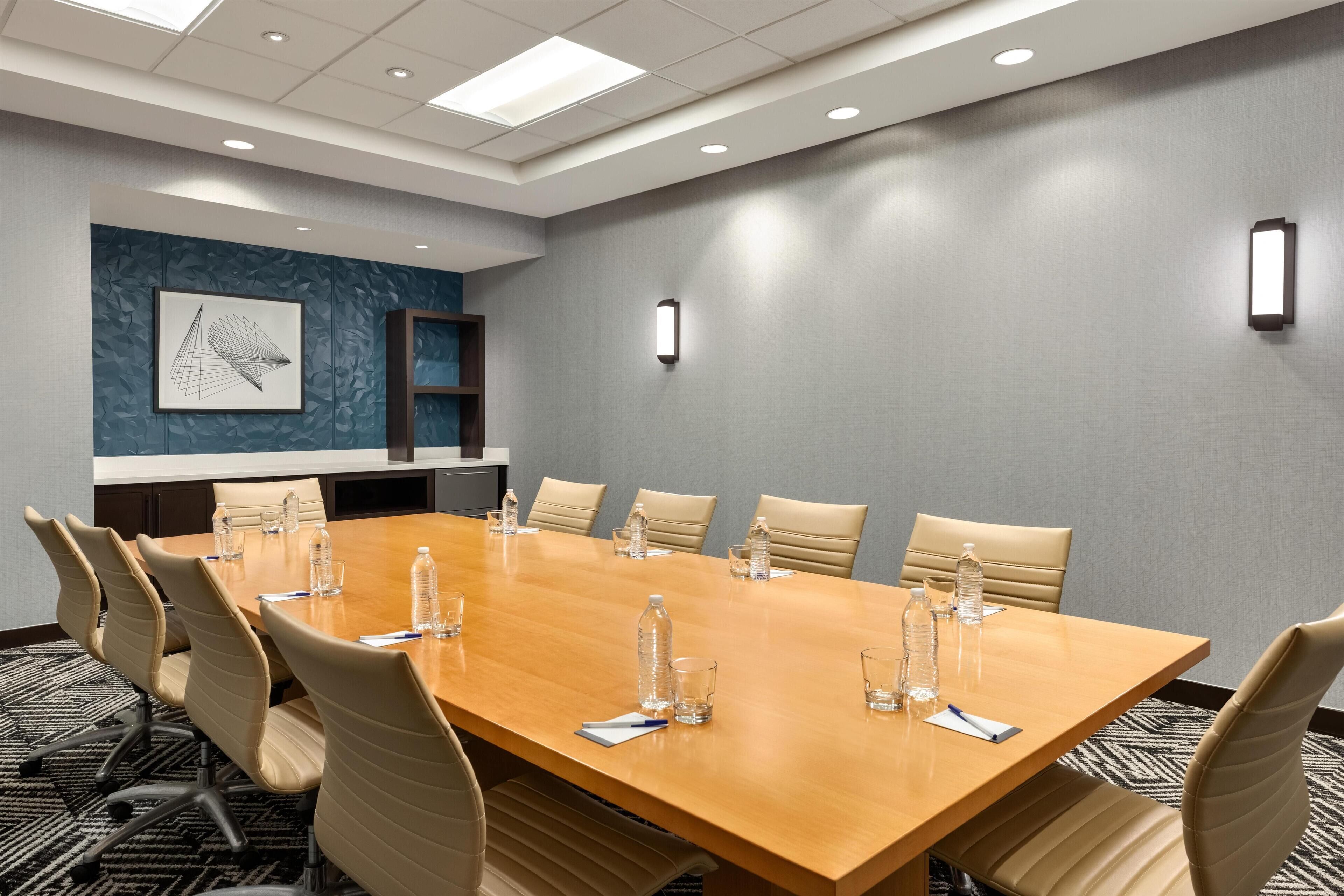 Un salón de conferencias con una mesa larga, sillas color beige, botellas de agua y una pared azul con diseño geométrico.