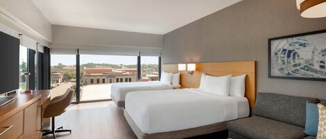Habitación de hotel con dos camas, un escritorio, un sofá y un amplio ventanal con vistas a la ciudad.