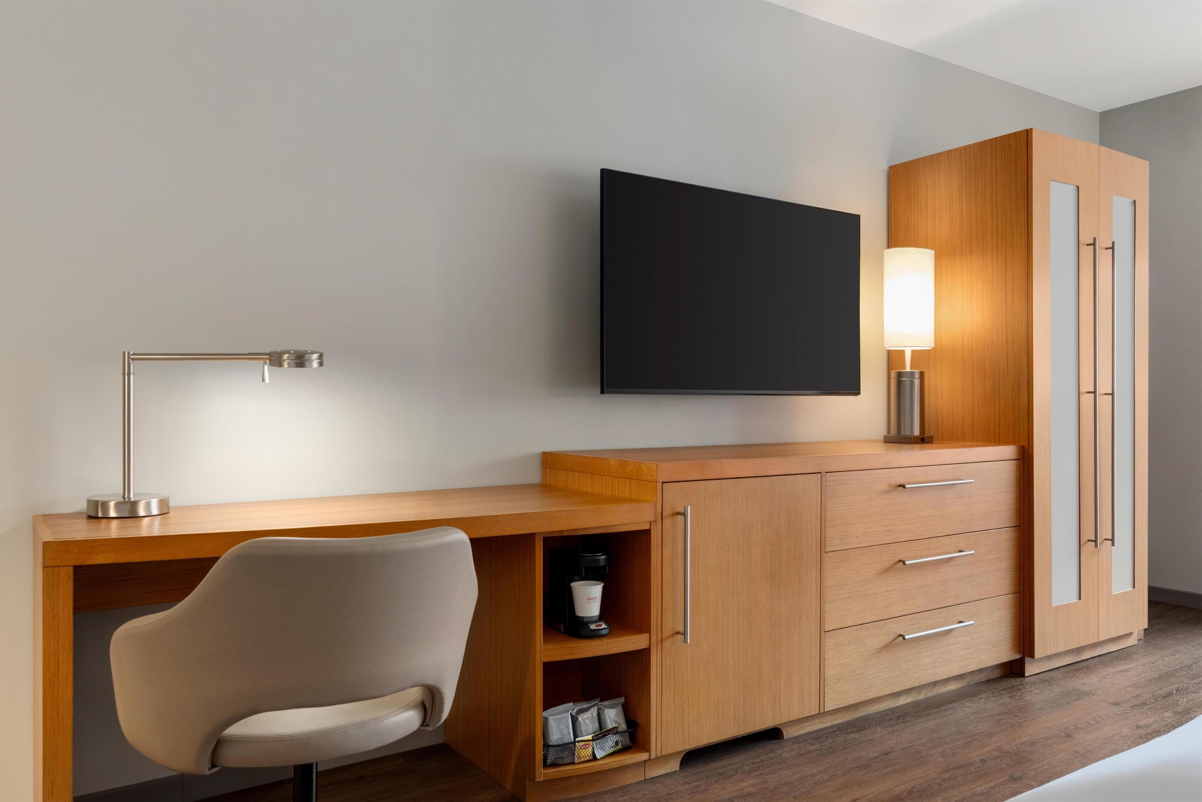 Et moderne hotelværelse med skrivebord i træ, en stol, et fladskærms-tv, en sengebordlampe og et klædeskab.