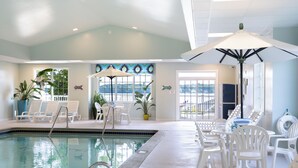 Une piscine intérieure à l’eau claire, entourée de chaises longues blanches et de parasols, et de grandes fenêtres offrant une vue sur l’extérieur.
