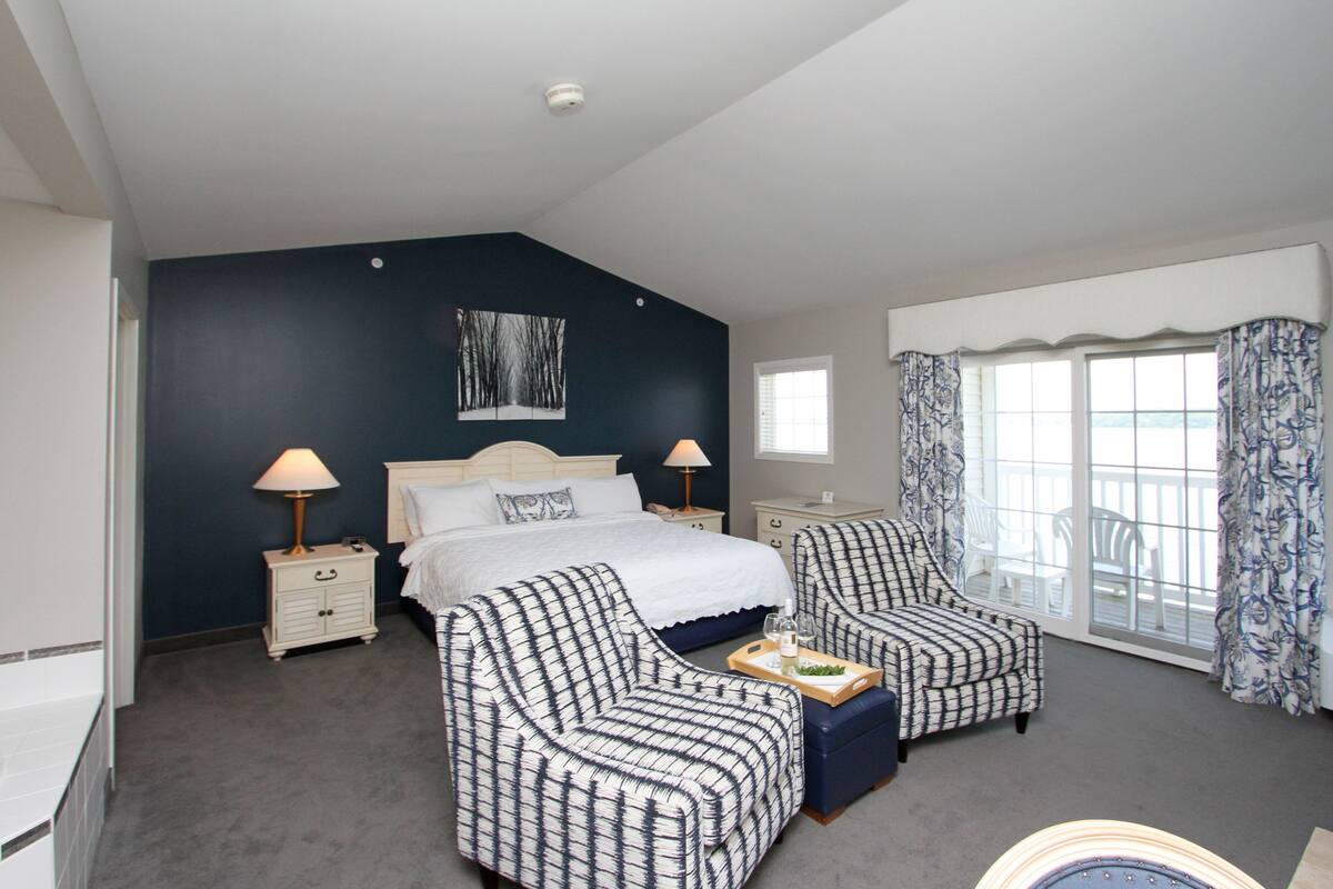 A bedroom with a bed, two bedside tables, a nightstand, a dresser, two patterned armchairs, a window with curtains, and a view of the outdoors.