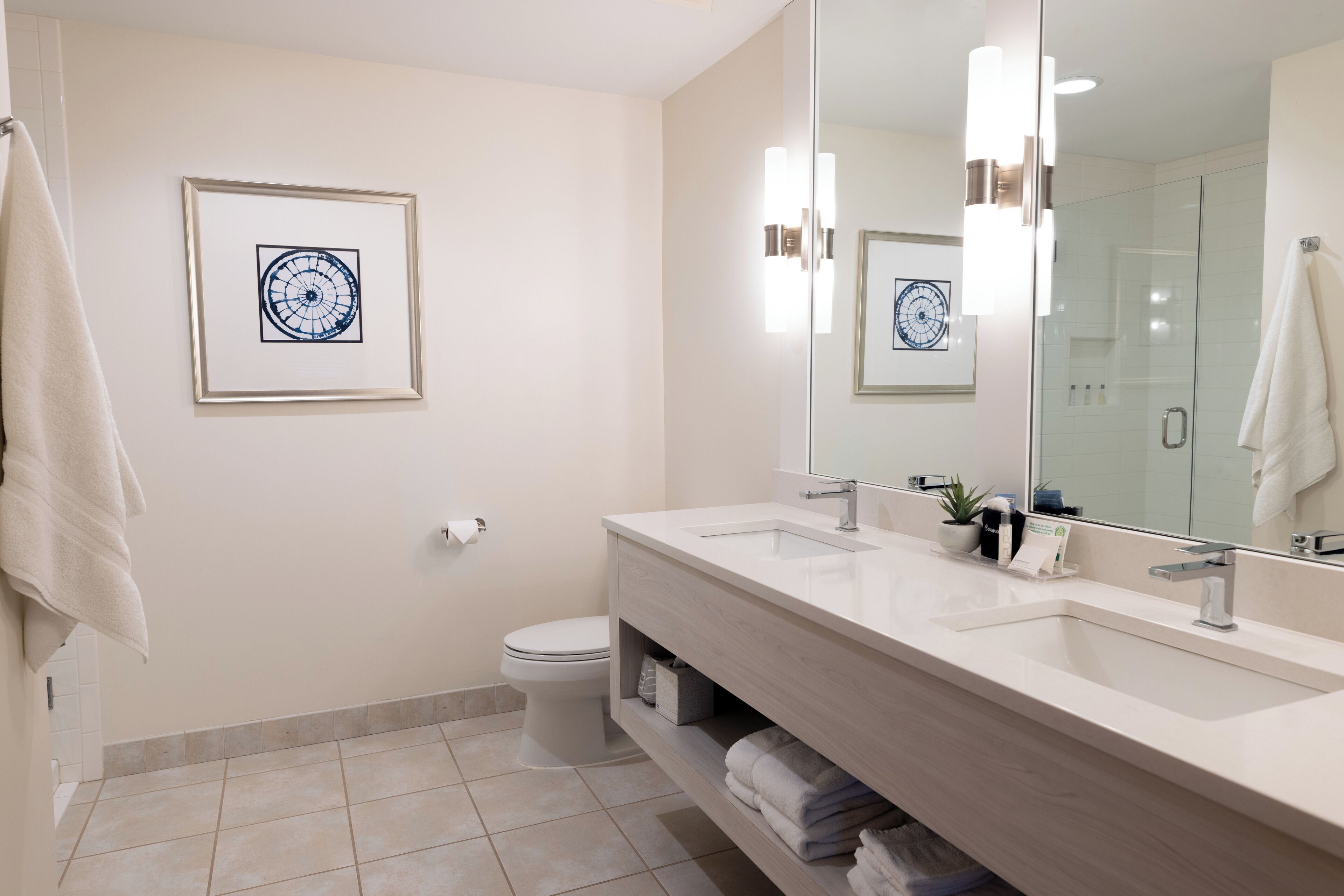 A modern bathroom with a large mirror, two sinks, a toilet, and a framed artwork on the wall.