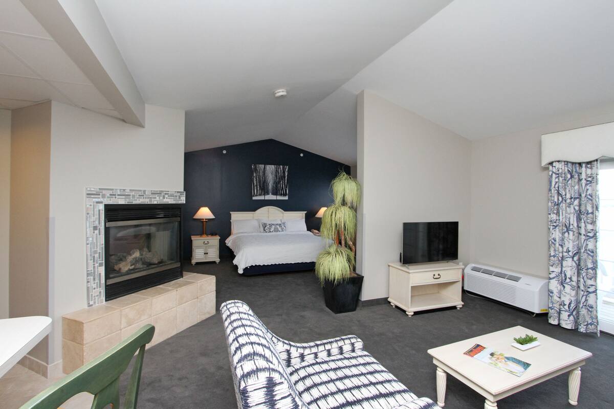 A modern hotel room with a fireplace, a bed with a headboard, a TV, a small table, and a potted plant.