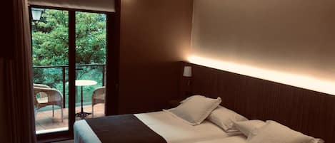 A hotel room with a bed, a nightstand, a lamp, and a view of a balcony with chairs and greenery.