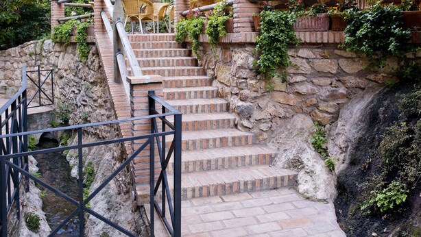 Gradians de pedra que condueixen a una zona d'aixetes amb mobles de vímet, envoltada de vegetació i un penya-segat rocos.