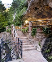 Una escalera de piedra conduce a una zona de estar con muebles de mimbre, rodeada de vegetación y un risco rocoso.