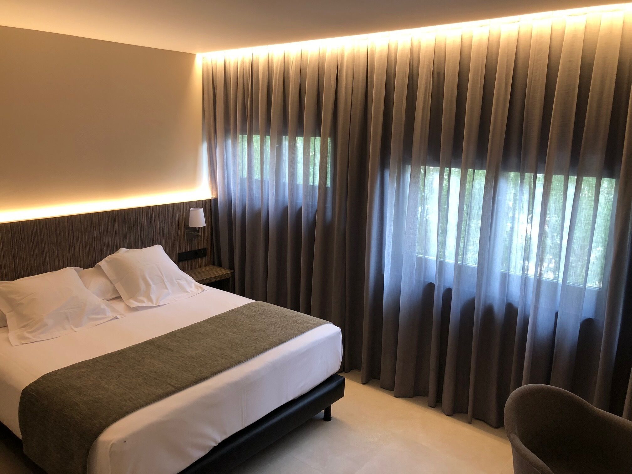 Una habitación de hotel con una cama grande, una mesita de noche con lámpara y una ventana con cortinas.