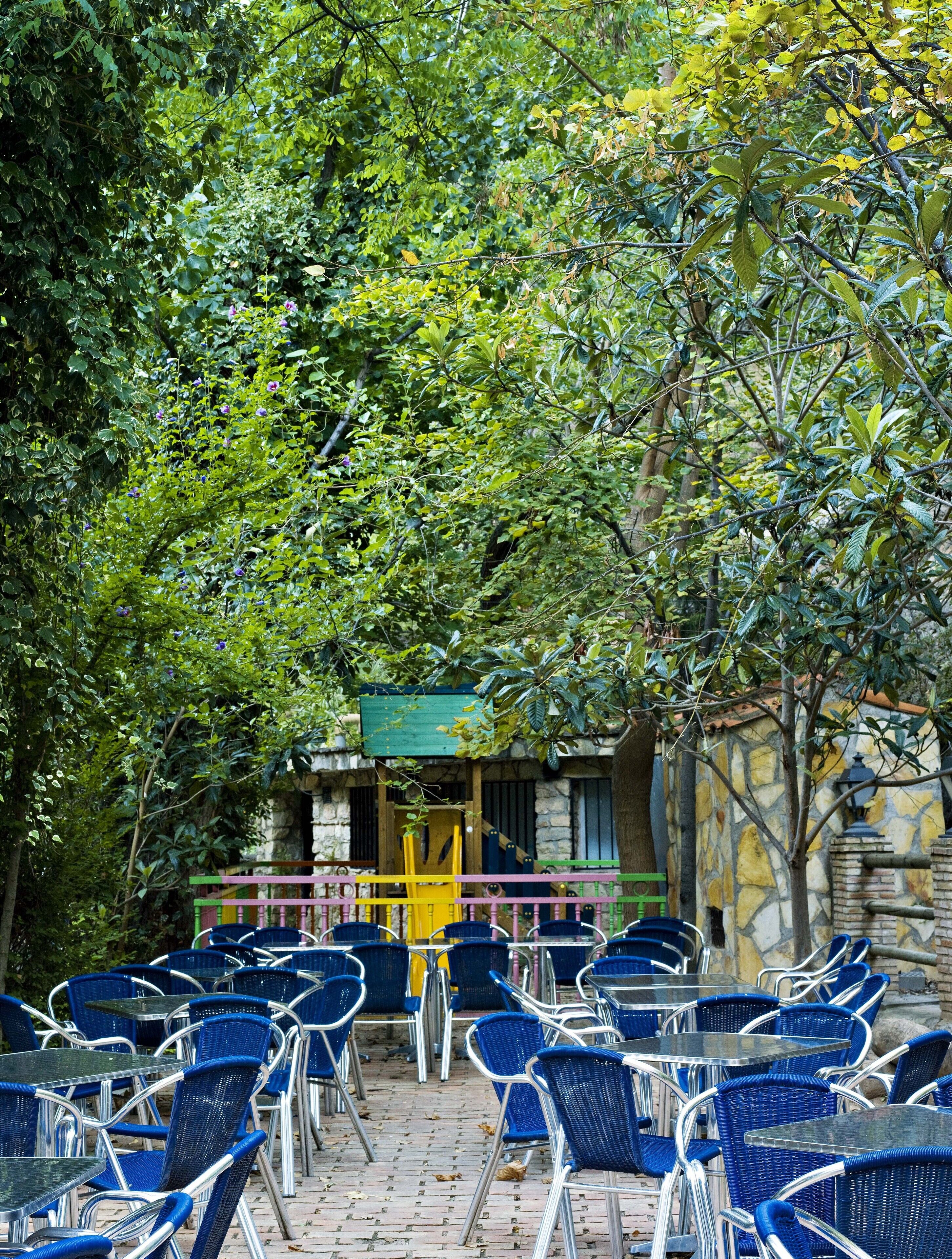 An outdoor seating area with blue chairs and tables, surrounded by lush greenery and a stone building with a colorful railing.