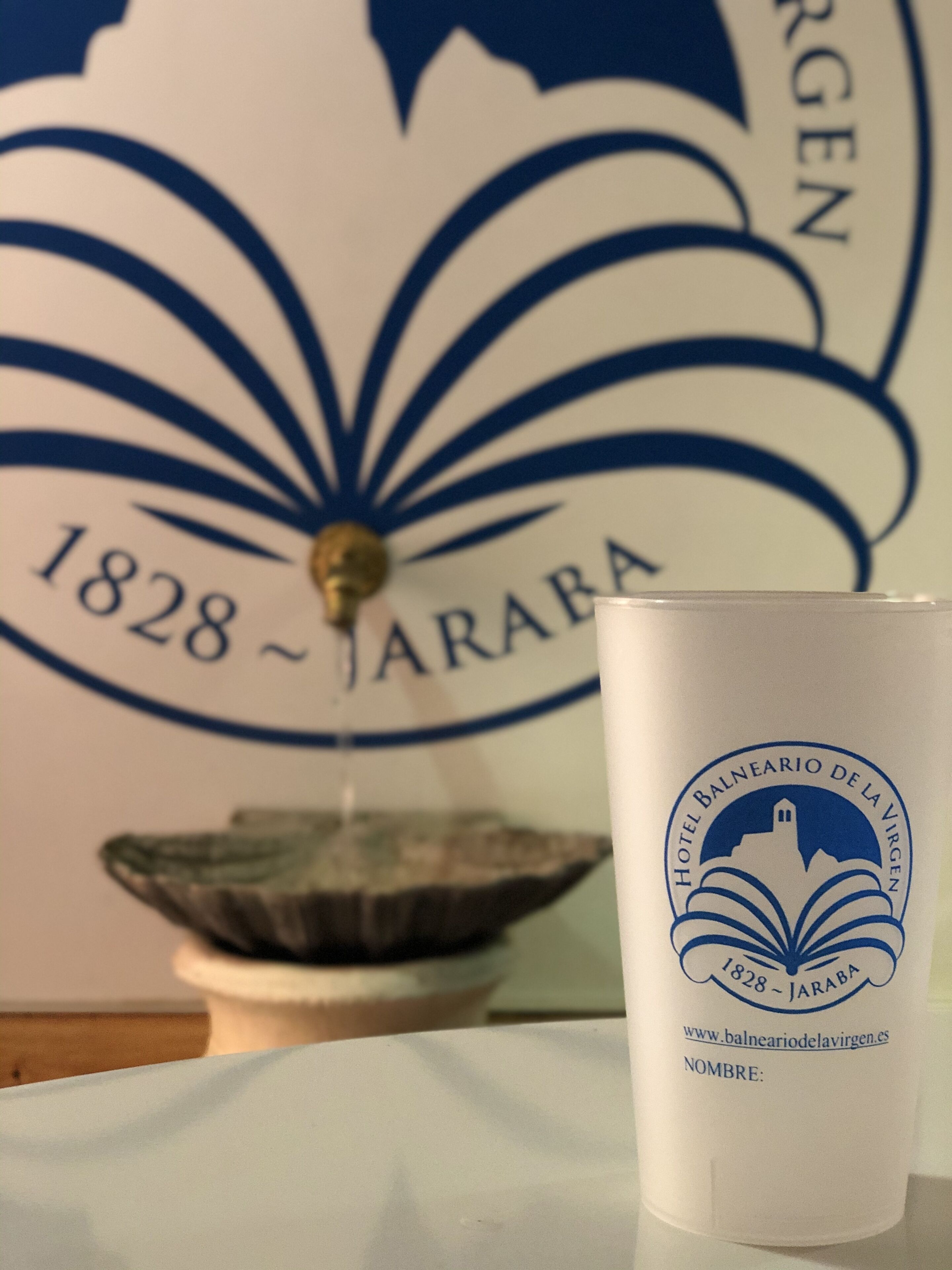 En vit mugg med en logotyp och text som visar att den kommer från Hotel Balneario de la Virgen, som grundades 1828 i JARABA.