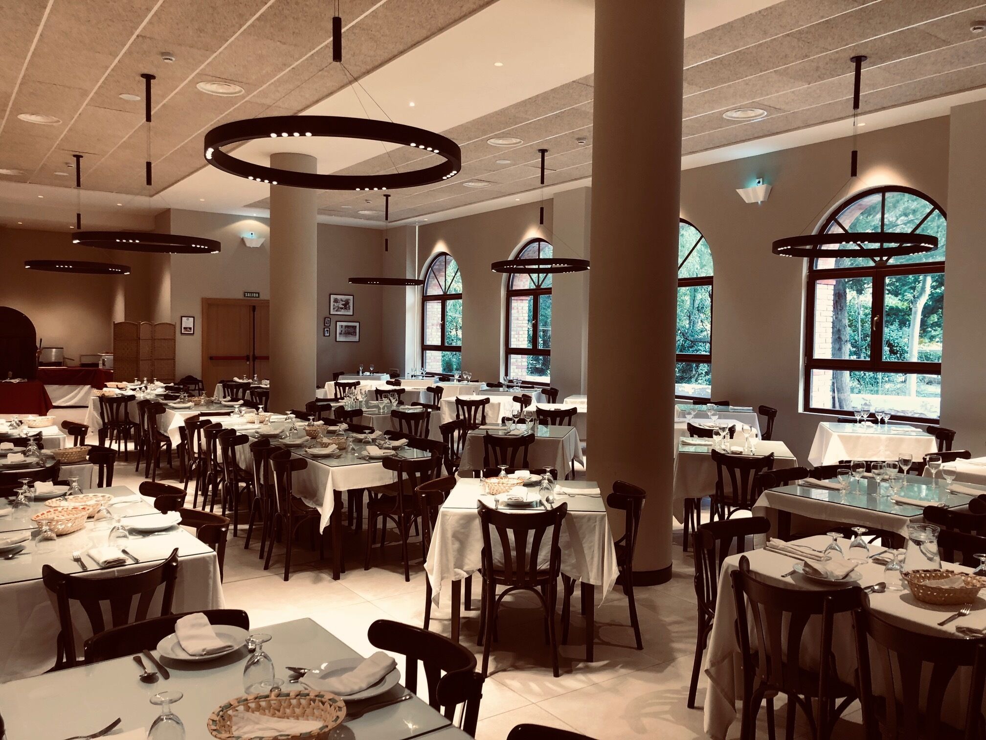 Un comedor amplio con varias mesas dispuestas para comer, ventanas grandes que permiten la entrada de luz natural e iluminación colgante moderna.