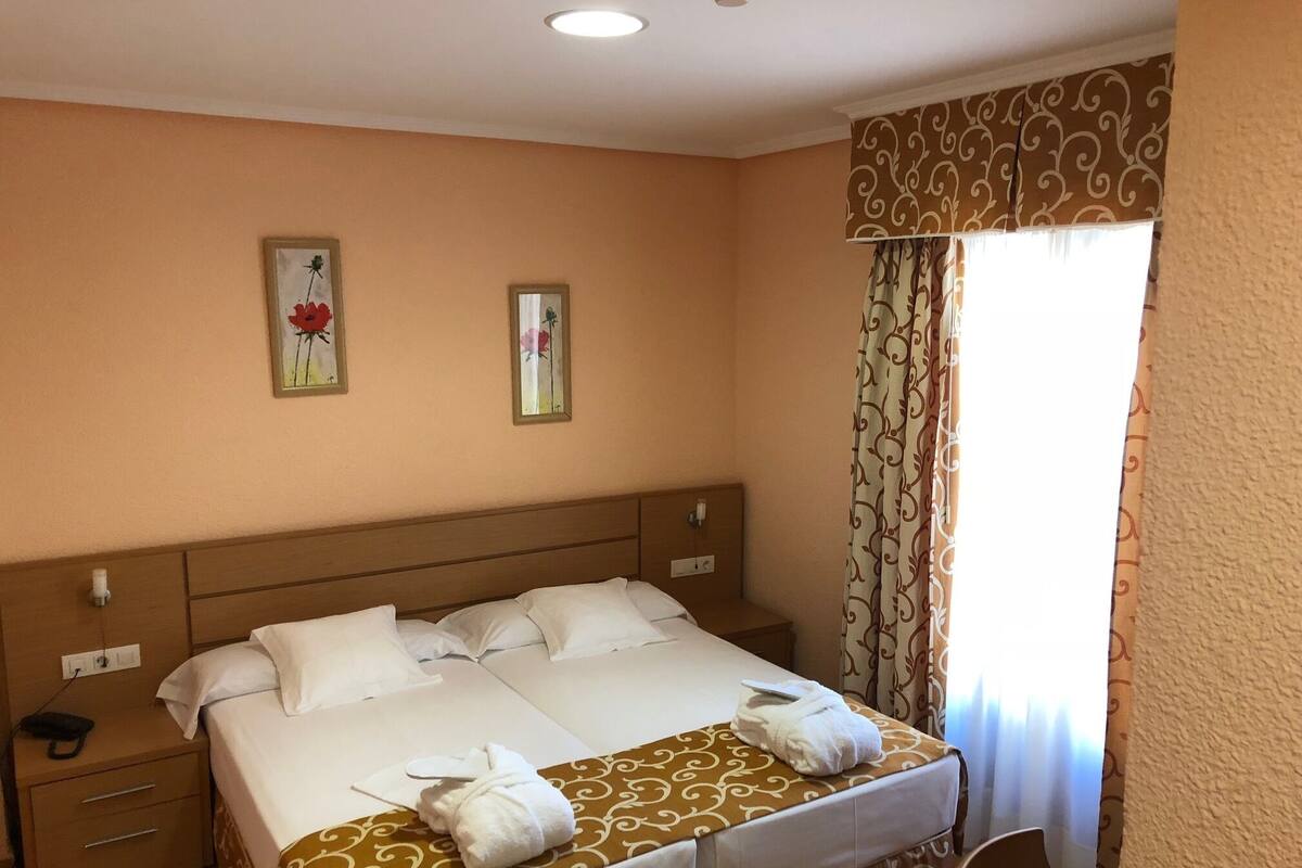 Una habitación de hotel con una cama grande, dos mesitas de noche y un ventanal con cortinas.