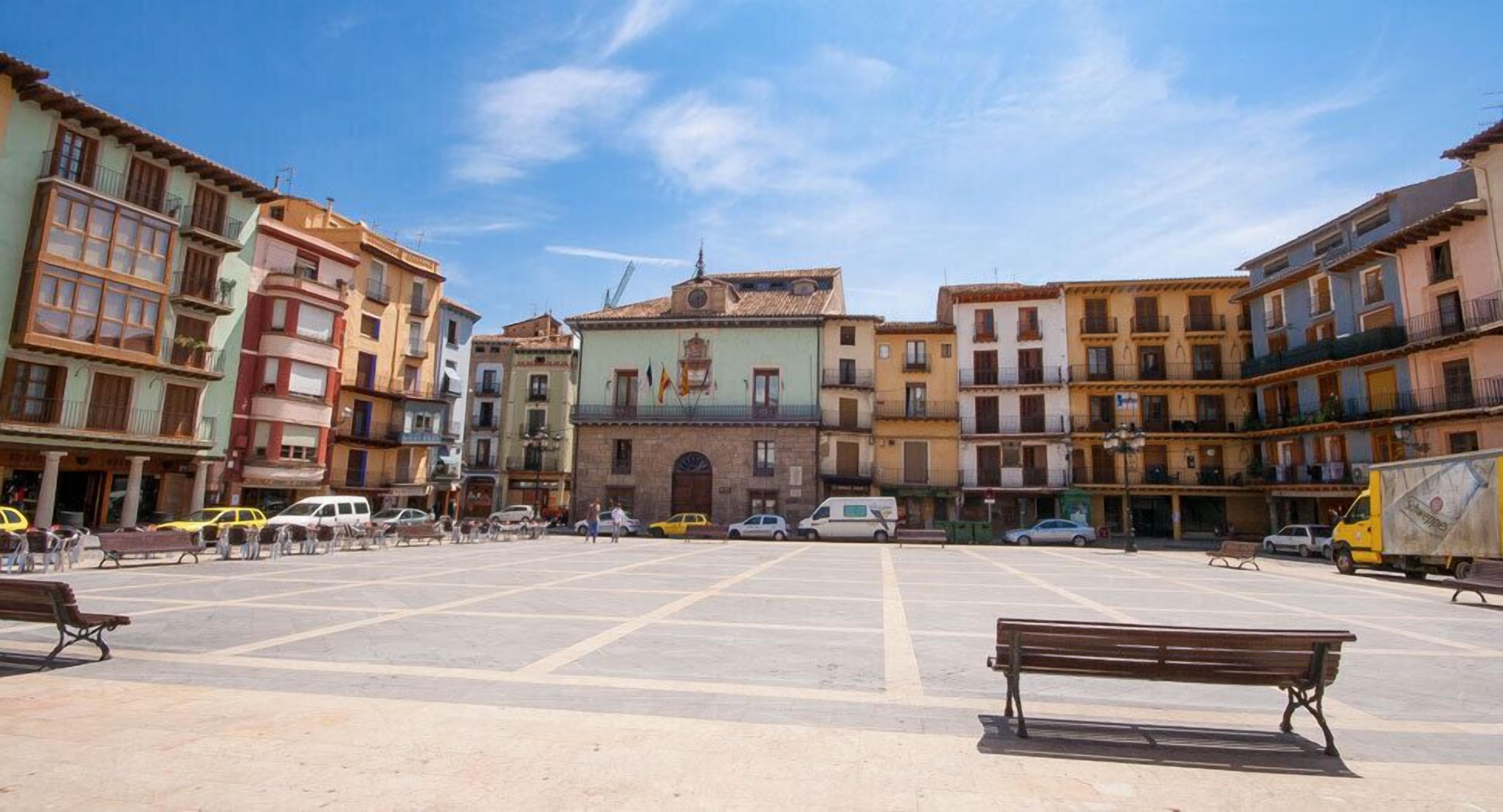 Una plaza con un edificio central, bancos y coches aparcados.