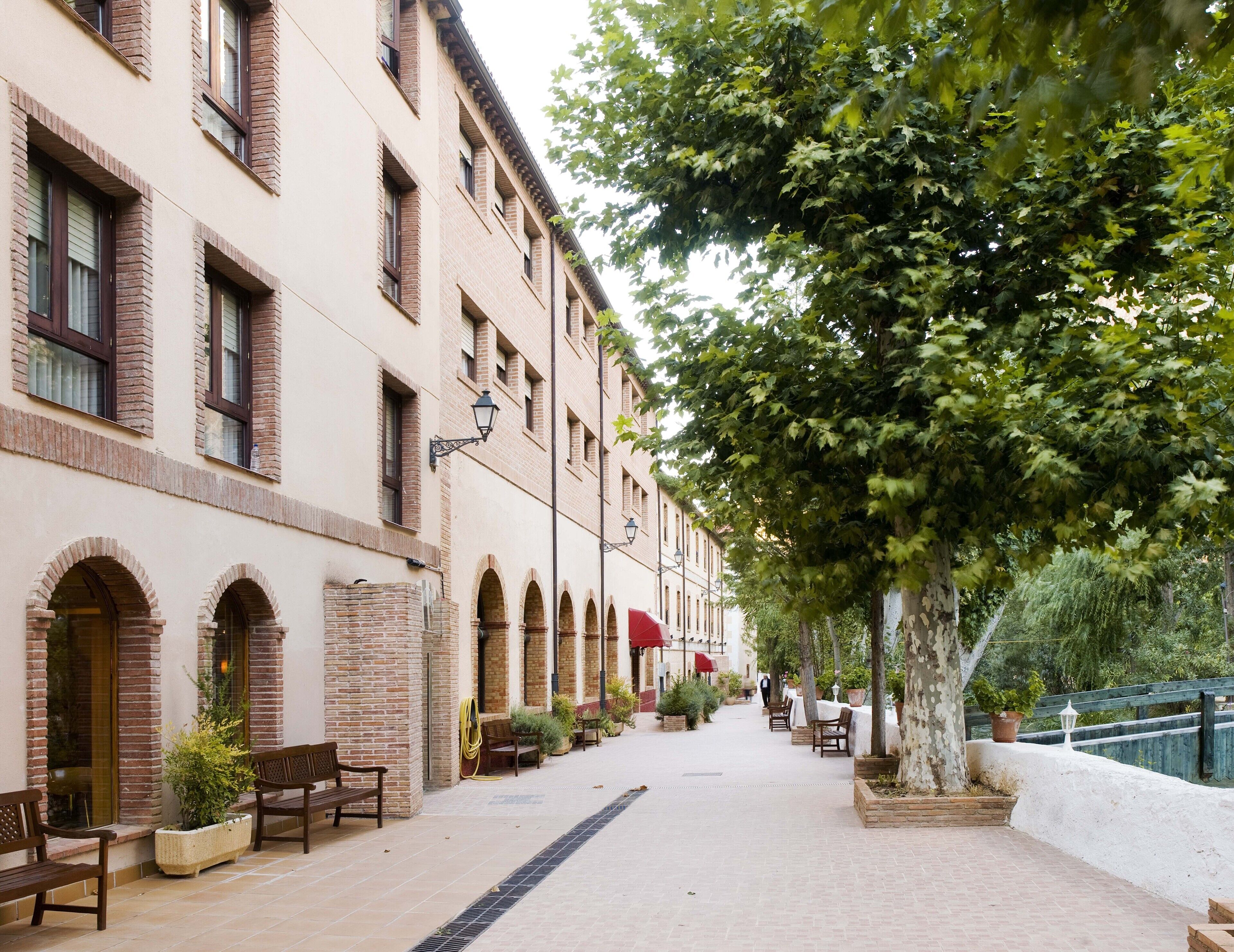 A building with multiple windows and arched doorways, a paved walkway, outdoor seating, and a tree providing shade.