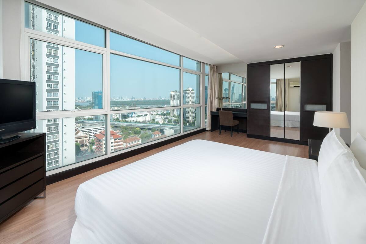 A modern hotel room with a large bed, a desk, a chair, a TV, and a view of the cityscape through large windows.