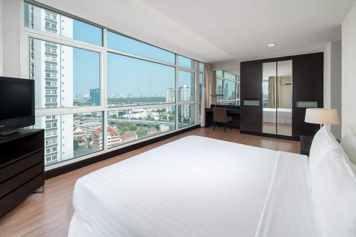 Ein modernes Hotelzimmer mit einem großen Bett, einem Schreibtisch, einem Sessel, einem Fernseher und Blick auf die Skyline durch große Fenster.