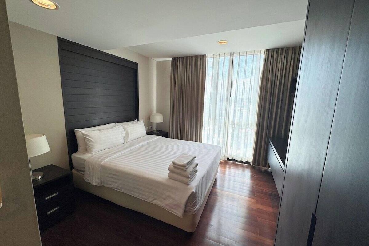 A modern hotel room with a large bed, dark headboard, bedside lamps, and a view of the city through large windows.