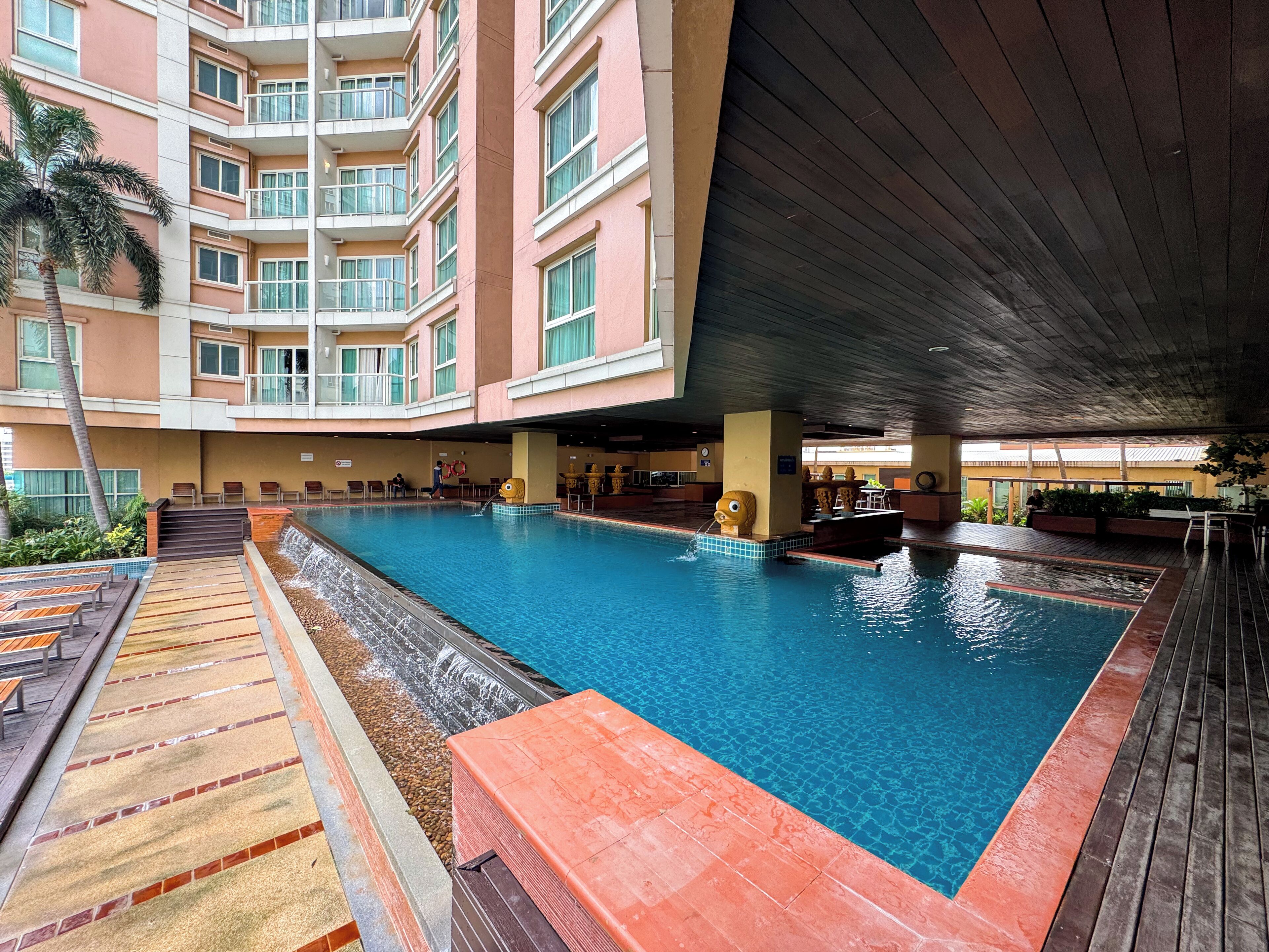 A swimming pool with a wooden deck, surrounded by a multi-story building with balconies.