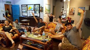 Eine Gruppe von Menschen sitzt auf gemusterten Sofas in einem Raum mit einem Holztisch, einer blauen Tür und einem Korkbrett mit verschiedenen Papieren und Fotos.