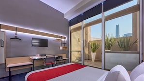Una habitación de hotel moderna con una cama grande, un escritorio con una silla, un televisor y un balcón con plantas.