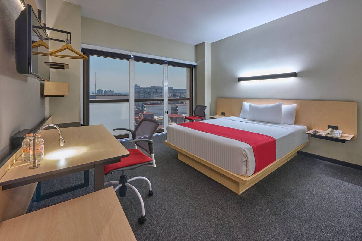 A hotel room with a bed, desk, chair, and a large window offering a city view.