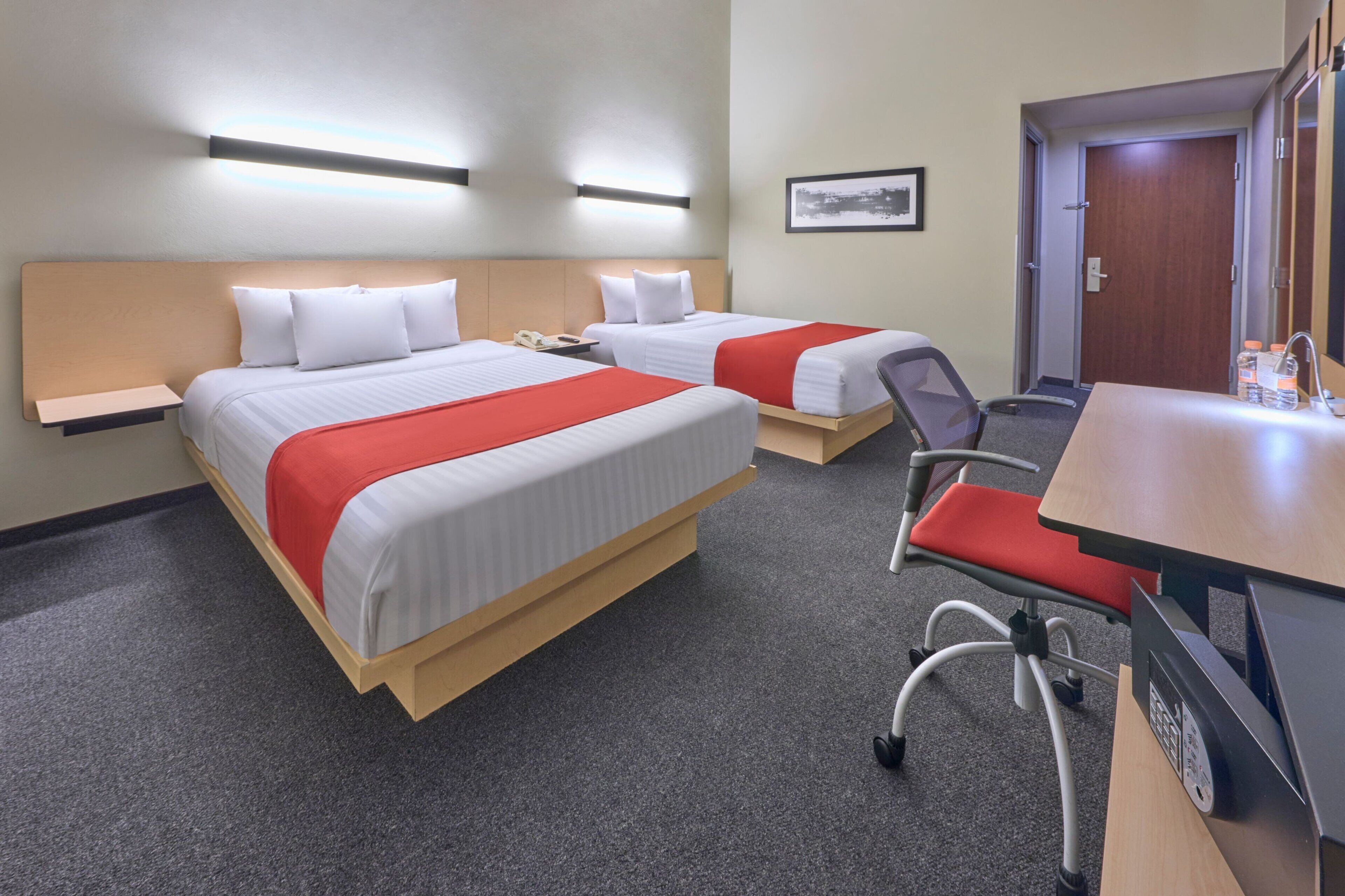 A hotel room with two beds, a desk, and a chair. There is a nightstand with a phone on each bed.