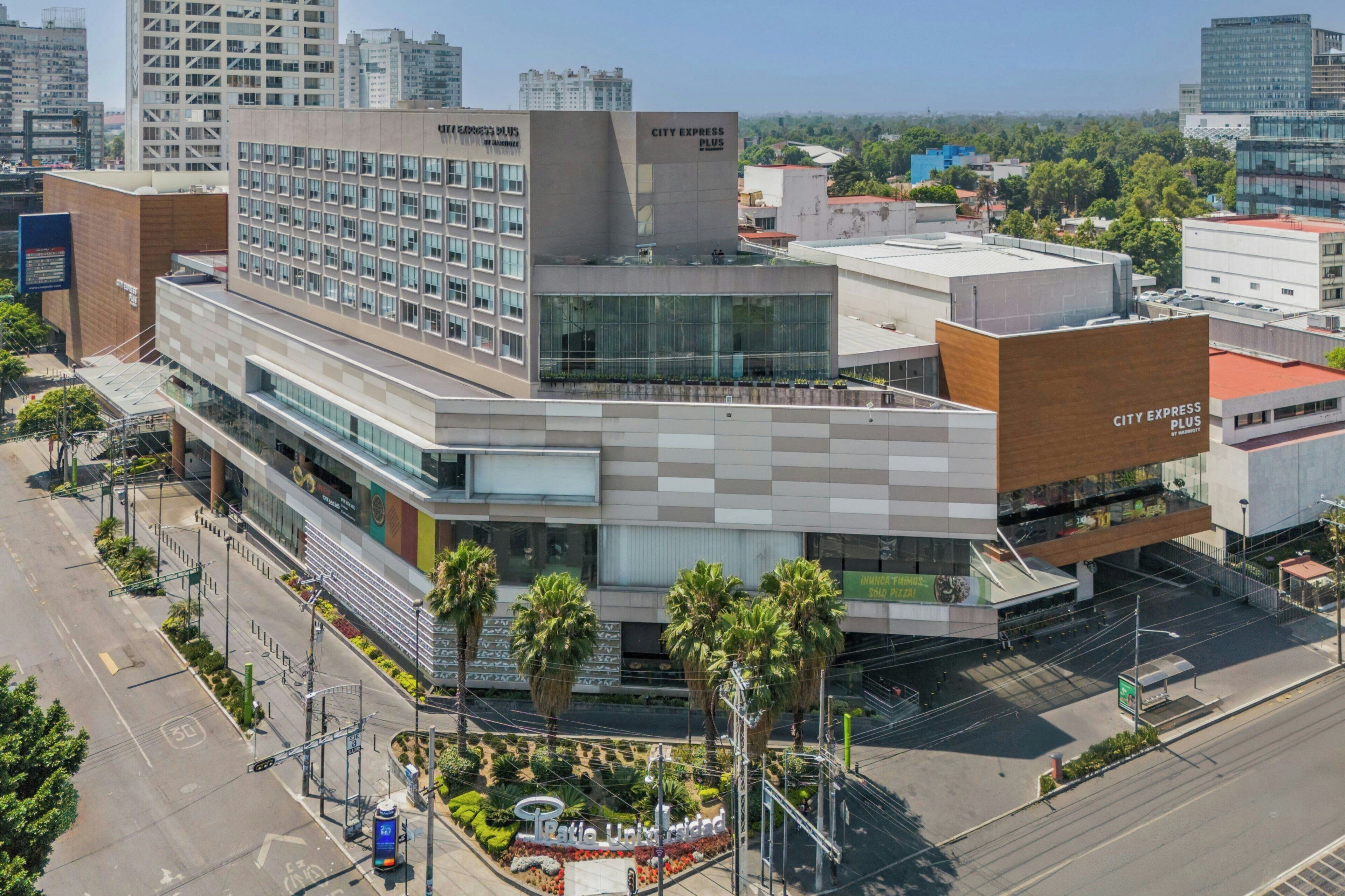 A modern building with a City Express Plus sign, a large glass facade, and a landscaped area with palm trees.