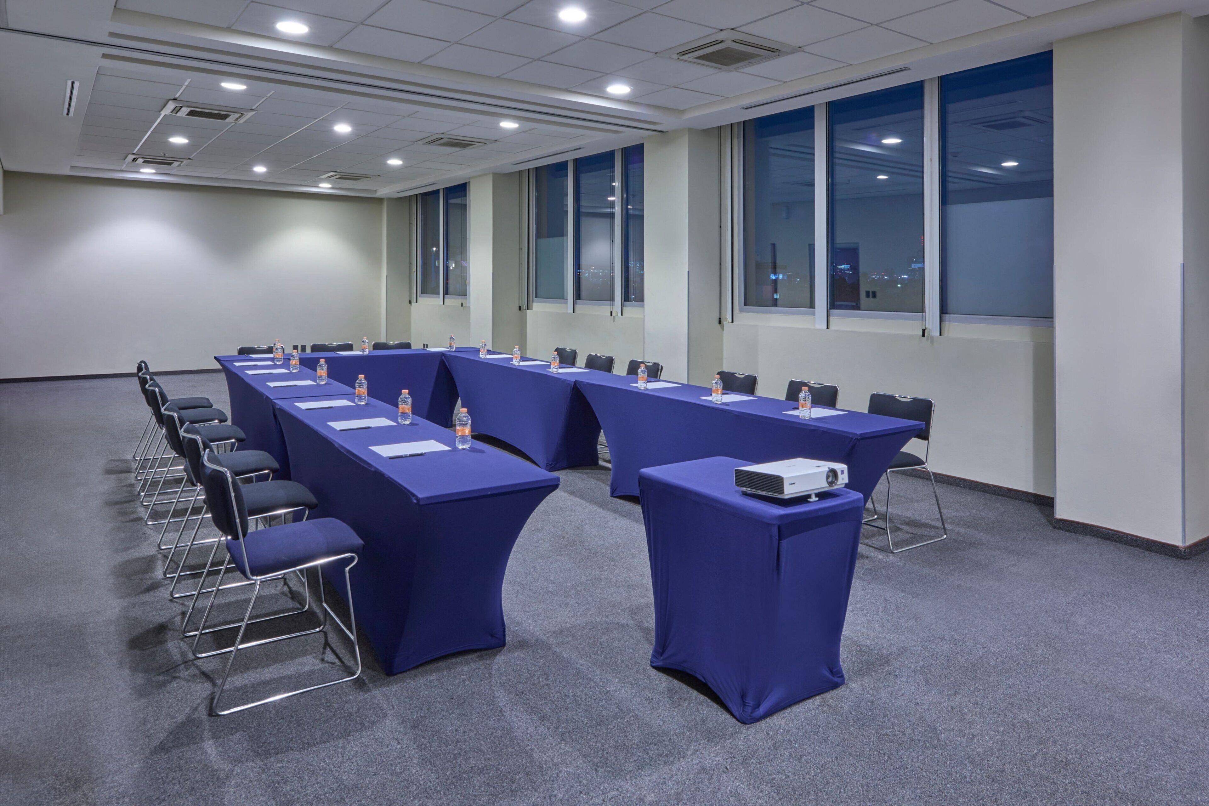 Un salón de conferencias con largas mesas, sillas y un proyector.