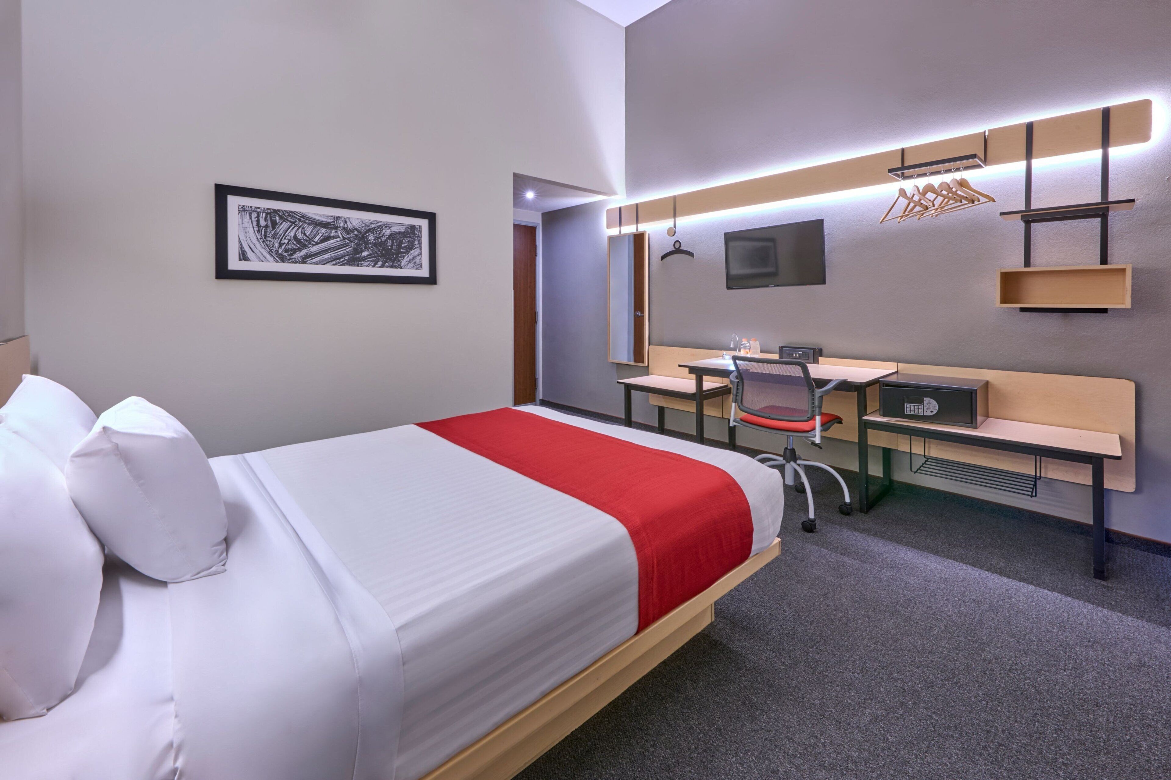 A hotel room with a bed, a desk, a chair, a TV, and a framed picture on the wall.