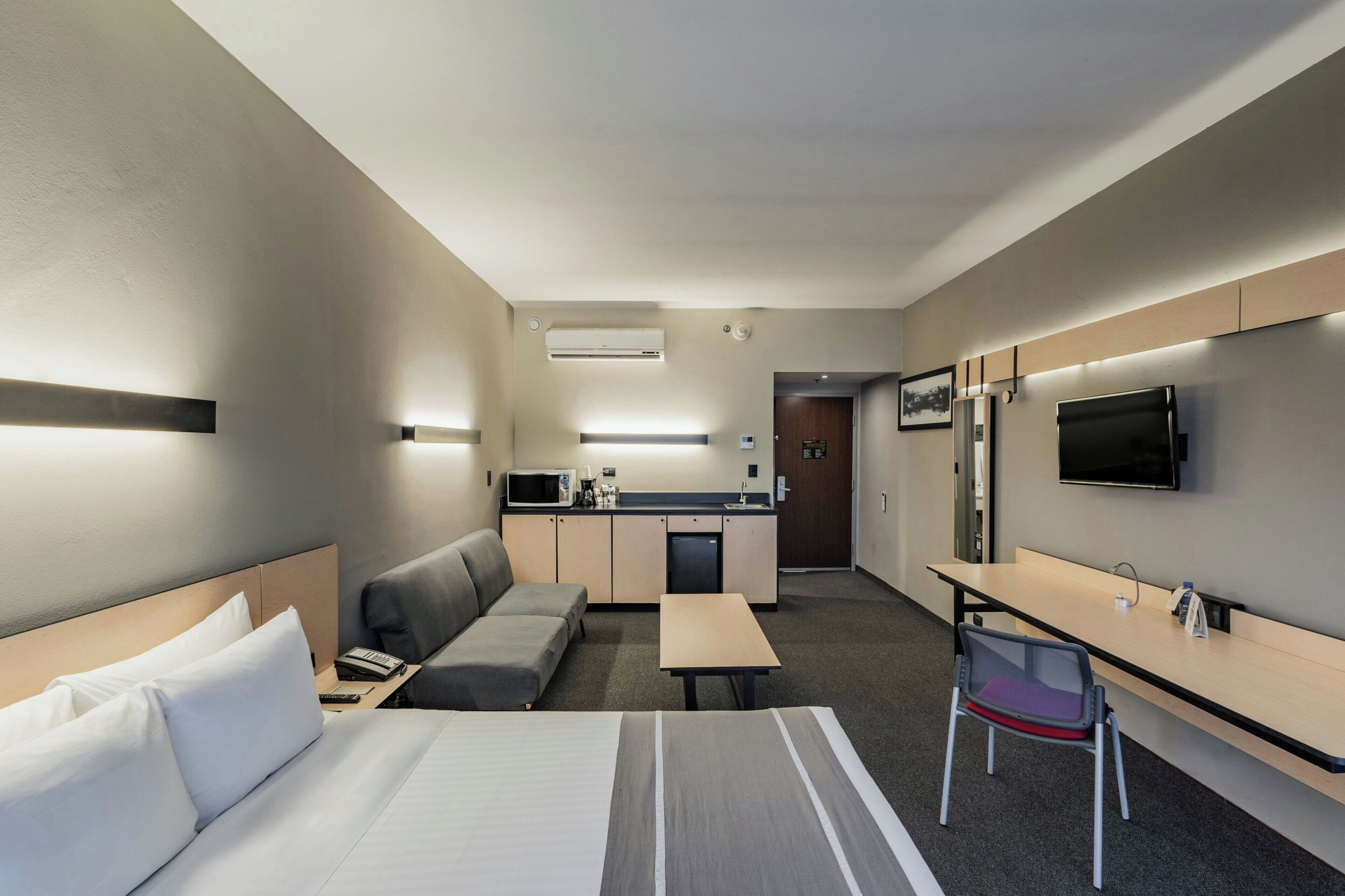 A hotel room with a bed, a sofa, a desk, a chair, a TV, and a kitchenette.