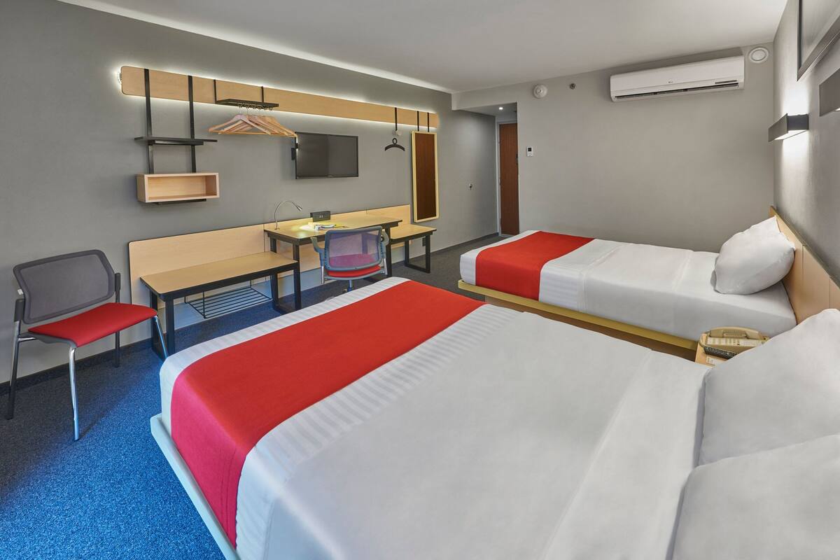 A hotel room with two beds, a desk, a chair, a TV, and an air conditioning unit.